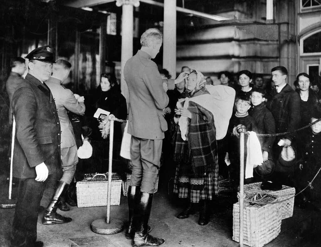 Image shows a male public health service officer checking an immigrant woman for signs of illness at Ellis Island, New York