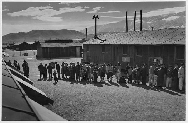 Image shows people of all ages lining up at Manzanar Camp in front of a mess hall