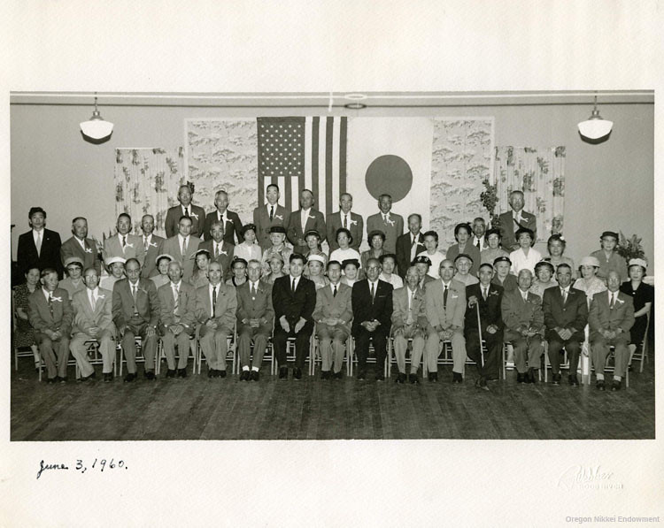 Image shows men and women seated in rows in front of an American and Japanese flag during a citizenship ceremony