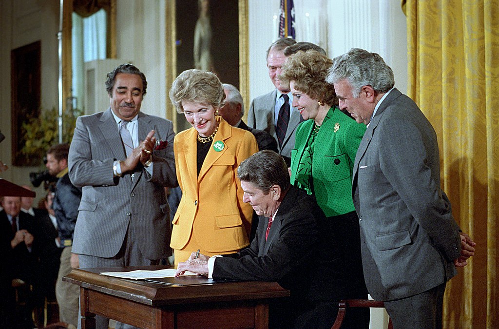 Image shows President Ronald Reagan sitting at a desk signing the Anti Drug Abuse Act of 1986, joined by the first lady, the vice president and senators. 