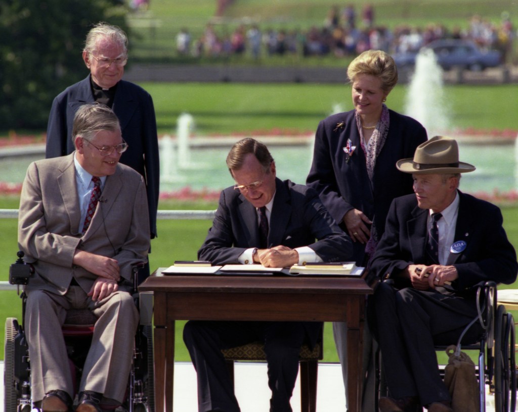 Image shows president George Bush signing the Americans with Disabilities Act (ADA) of 1990 at the White House, surrounded by ADA activists 
