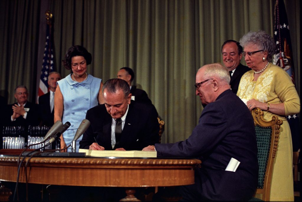 Image shows President Lyndon B. Johnson signing the Medicare bill surrounded by a former president, vice presidents, first lady, and commisioners