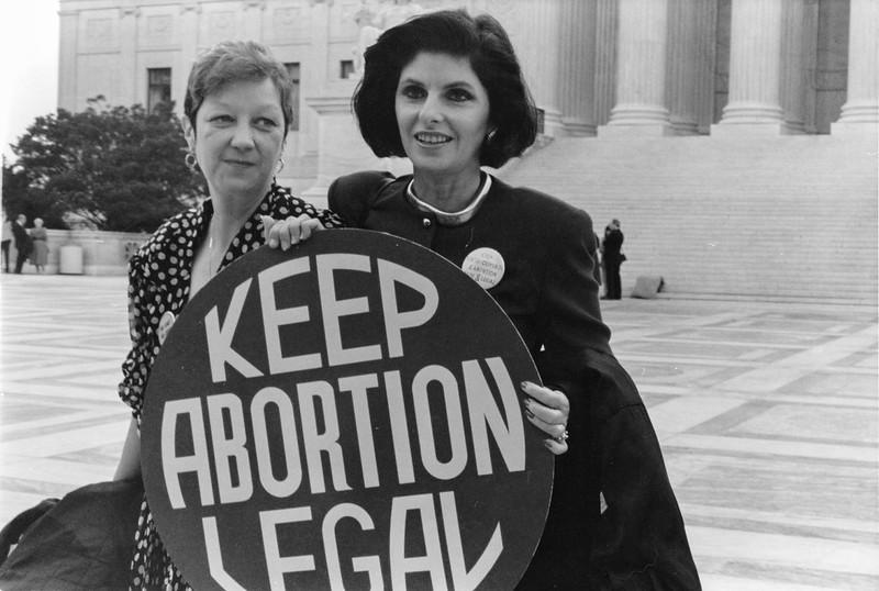 Image shows Norma McCorvey, known as Jane Roe, and her attorney holding a "Keep abortion legal" sign outside of the Supreme Court