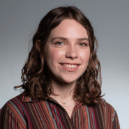 Photo of Abby Sachar in an red-earth-toned striped button down against a gray background