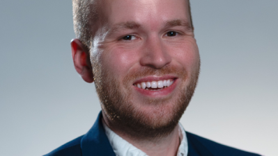Photo of Joel Luther in a white button up with a black blazer against a gray background.