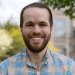 Photo of Joel Luther in a plaid shirt with trees and a building in the background