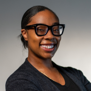 Headshot photo of Natiya Clark in a black top and black glasses against a gray background.