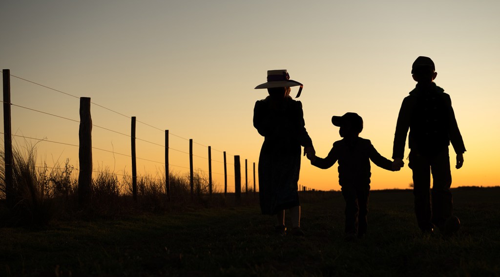 Shadowed figures of two parents and a young child walk along a fenceline holding hands at sunset.