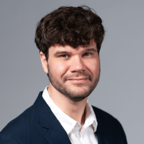 Professional headshot of Andrew Jones wearing a white collared shirt with a blue suit jacket against a gray background.