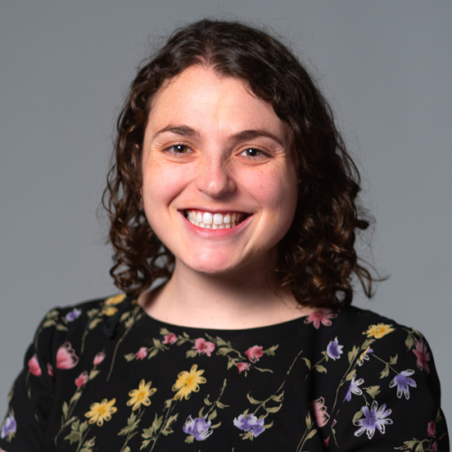 Professional headshot of Maia Rosenfeld wearing a floral top against a gray background.