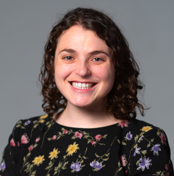 Professional headshot of Maia Rosenfeld wearing a floral top against a gray background.