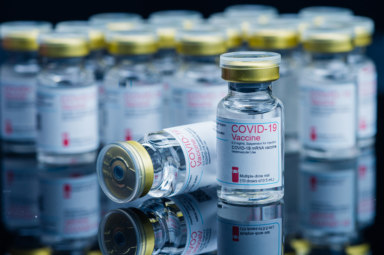 A stockpile of COVID vaccine vials on a flat, black surface is pictured with one tipped over in the foreground of the photo.