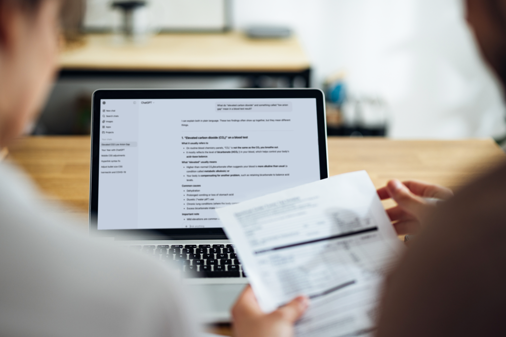 A laptop screen screen displaying a list is viewed over the shoulders of two people comparing it to a paper bill held in hand.