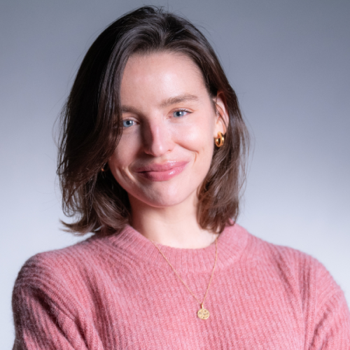 Professional headshot of Kate Wells in a light pink sweater against a gray background.