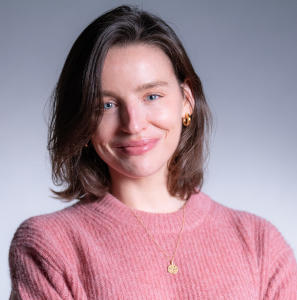 Professional headshot of Kate Wells in a light pink sweater against a gray background.