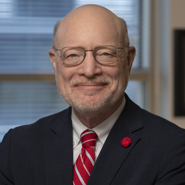 Headshot photo of Chip Kahn wearing a navy blue suit with a red tie, red pendant on lapel, and glasses.