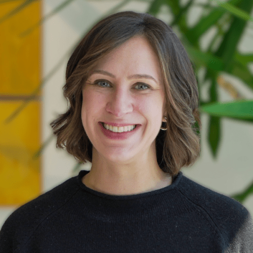 Professional headshot of Emily Hague in a black sweater against a sun-soaked wall.