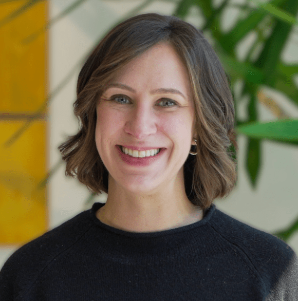 Professional headshot of Emily Hague in a black sweater against a sun-soaked wall.